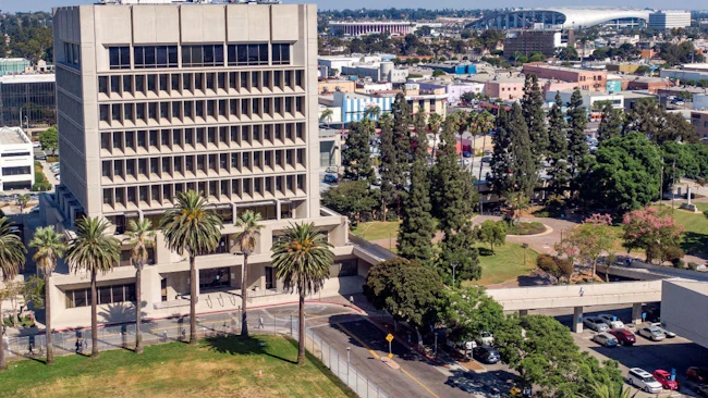 An aerial view of a mid-century concrete office tower in Downtown Inglewood, Calif., surrounded by palm trees and green space, with the broader Inglewood urban landscape, retail corridors, and a large stadium structure visible in the background.