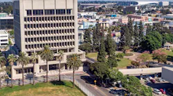 An aerial view of a mid-century concrete office tower in Downtown Inglewood, Calif., surrounded by palm trees and green space, with the broader Inglewood urban landscape, retail corridors, and a large stadium structure visible in the background.