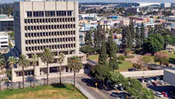 An aerial view of a mid-century concrete office tower in Downtown Inglewood, Calif., surrounded by palm trees and green space, with the broader Inglewood urban landscape, retail corridors, and a large stadium structure visible in the background.