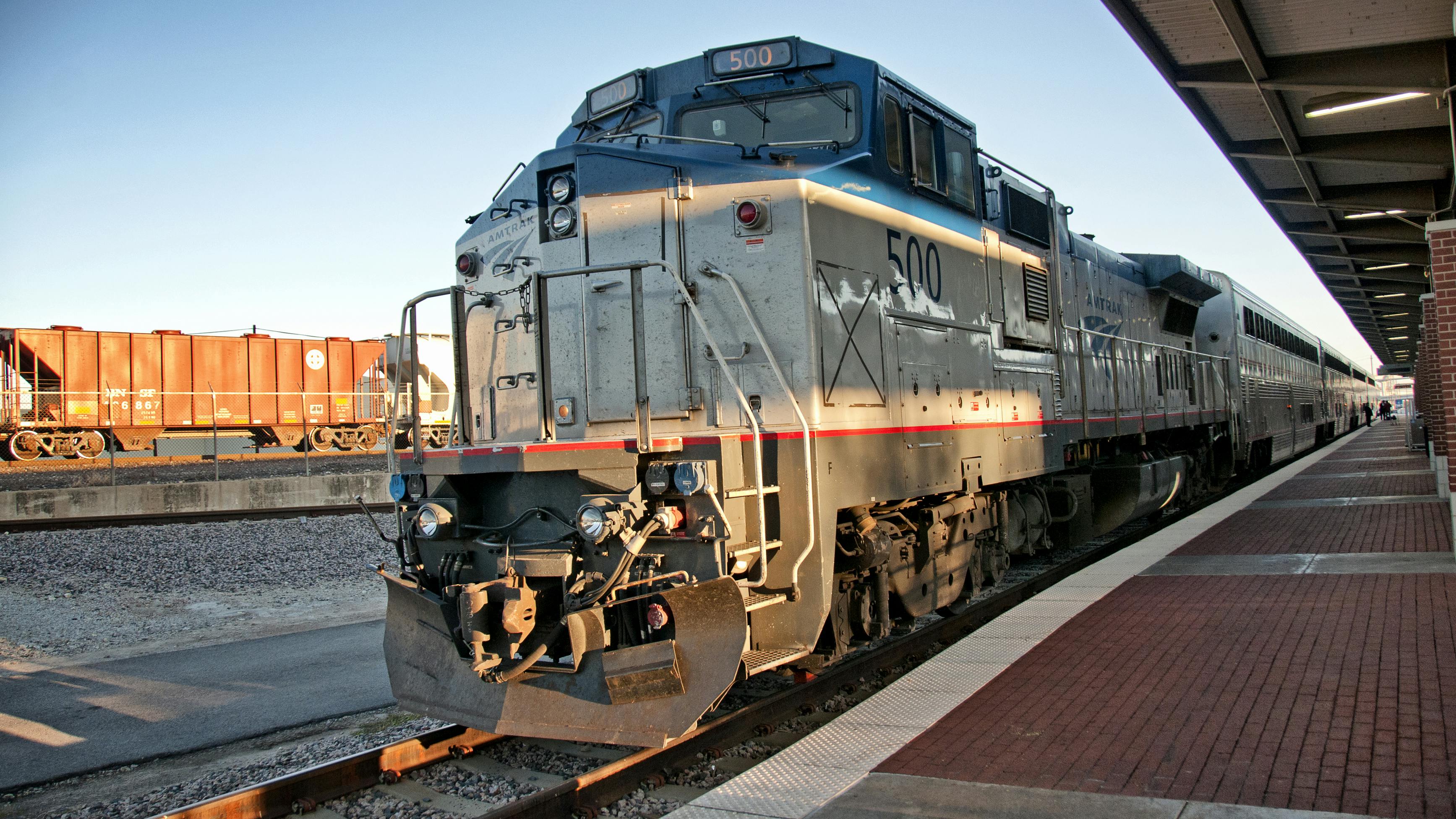 An Amtrak locomotive, numbered 500, sits at a station platform beside a freight train, with passenger cars trailing behind under a covered canopy at sunset.