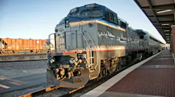 An Amtrak locomotive, numbered 500, sits at a station platform beside a freight train, with passenger cars trailing behind under a covered canopy at sunset.