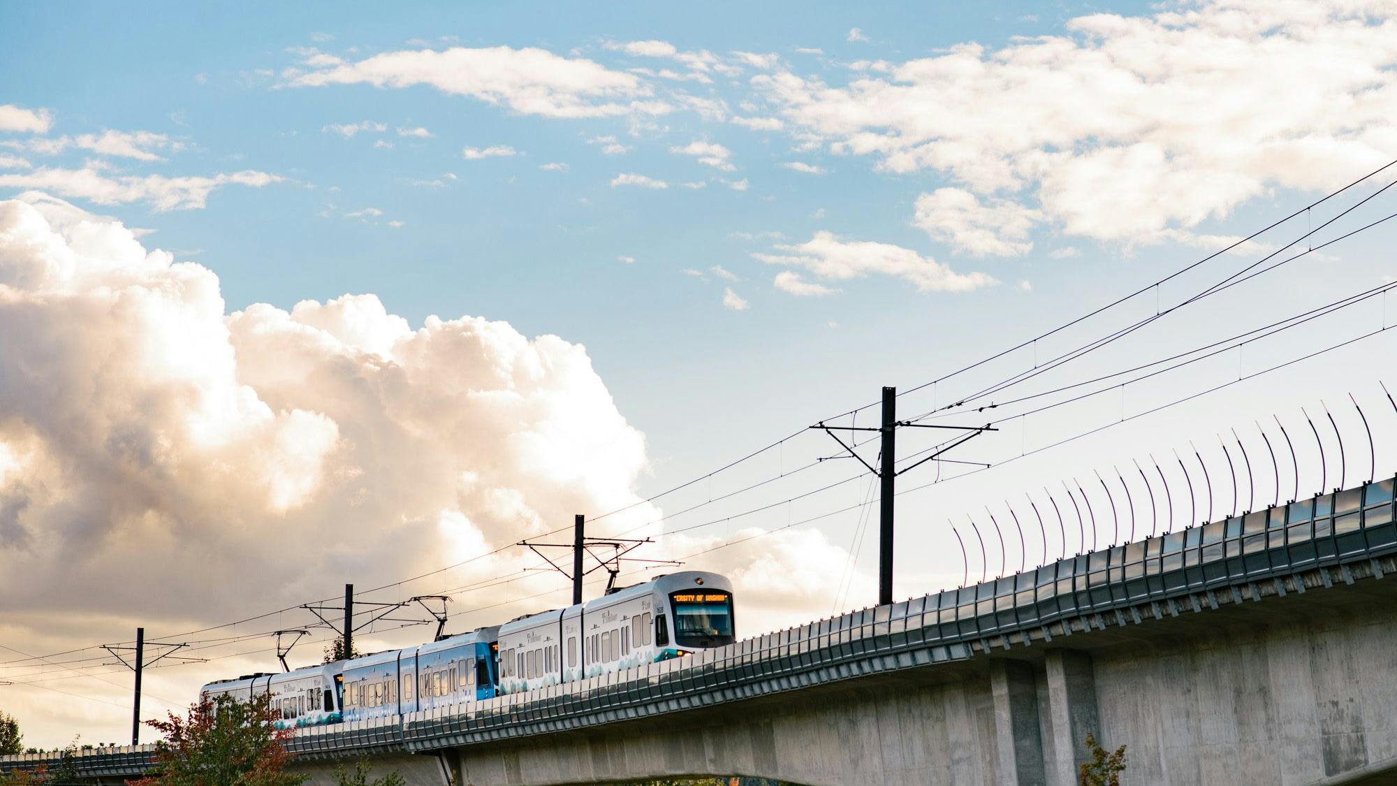 A Sound Transit Link light-rail train in white and teal livery travels across an elevated concrete guideway against a dramatic partly cloudy sky, with catenary wire infrastructure overhead and autumn-tinged trees visible below the viaduct.