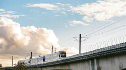 A Sound Transit Link light-rail train in white and teal livery travels across an elevated concrete guideway against a dramatic partly cloudy sky, with catenary wire infrastructure overhead and autumn-tinged trees visible below the viaduct.