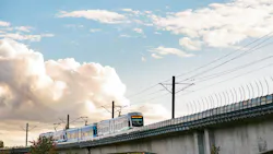 A Sound Transit Link light-rail train in white and teal livery travels across an elevated concrete guideway against a dramatic partly cloudy sky, with catenary wire infrastructure overhead and autumn-tinged trees visible below the viaduct.