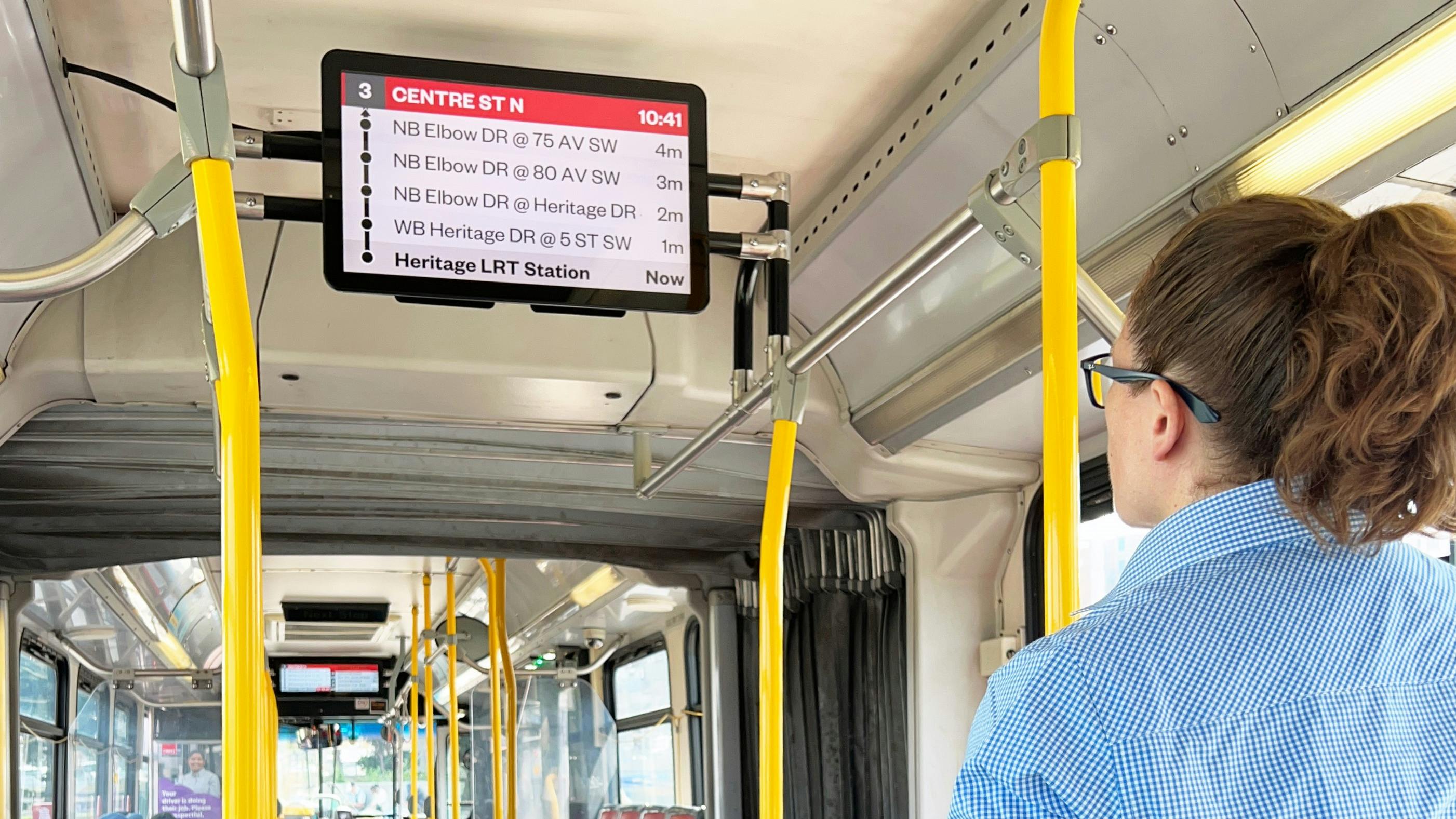 A Calgary Transit rider looks up at a new onboard passenger information display showing upcoming stops and arrival times for Route 3 Centre Street North, with the bus interior and other riders visible in the background.