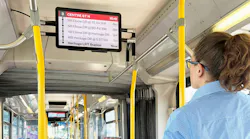 A Calgary Transit rider looks up at a new onboard passenger information display showing upcoming stops and arrival times for Route 3 Centre Street North, with the bus interior and other riders visible in the background.