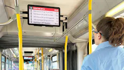 A Calgary Transit rider looks up at a new onboard passenger information display showing upcoming stops and arrival times for Route 3 Centre Street North, with the bus interior and other riders visible in the background.