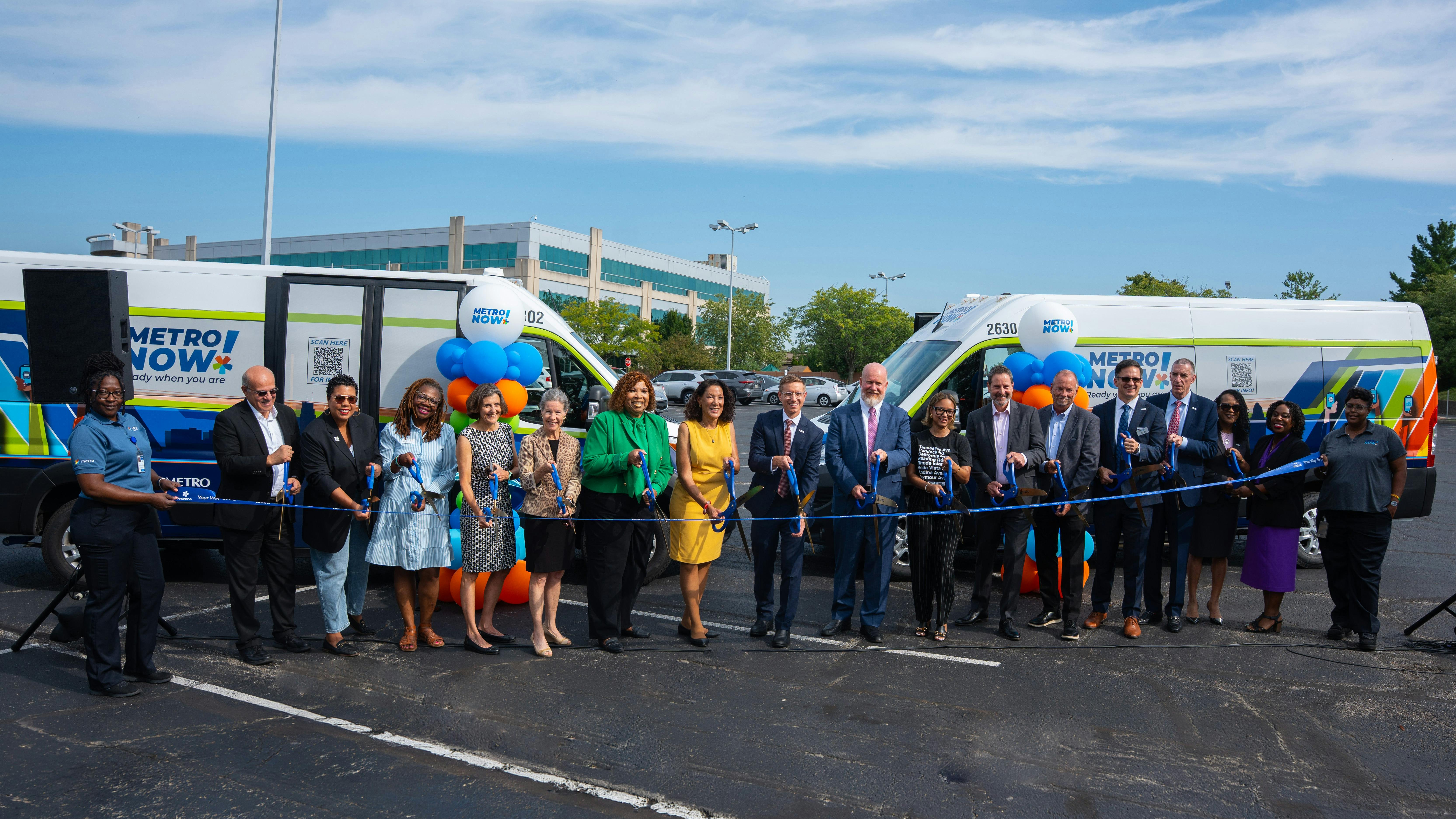 A large group of officials and Metro staff cut a ribbon in front of two 'Metro Now' microtransit vans wrapped in the service's branding, celebrating the launch of the Bond Hill-Roselawn service in a parking lot.