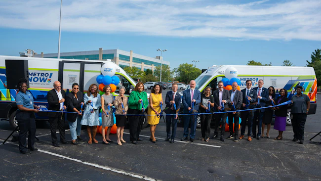 A large group of officials and Metro staff cut a ribbon in front of two 'Metro Now' microtransit vans wrapped in the service's branding, celebrating the launch of the Bond Hill-Roselawn service in a parking lot.