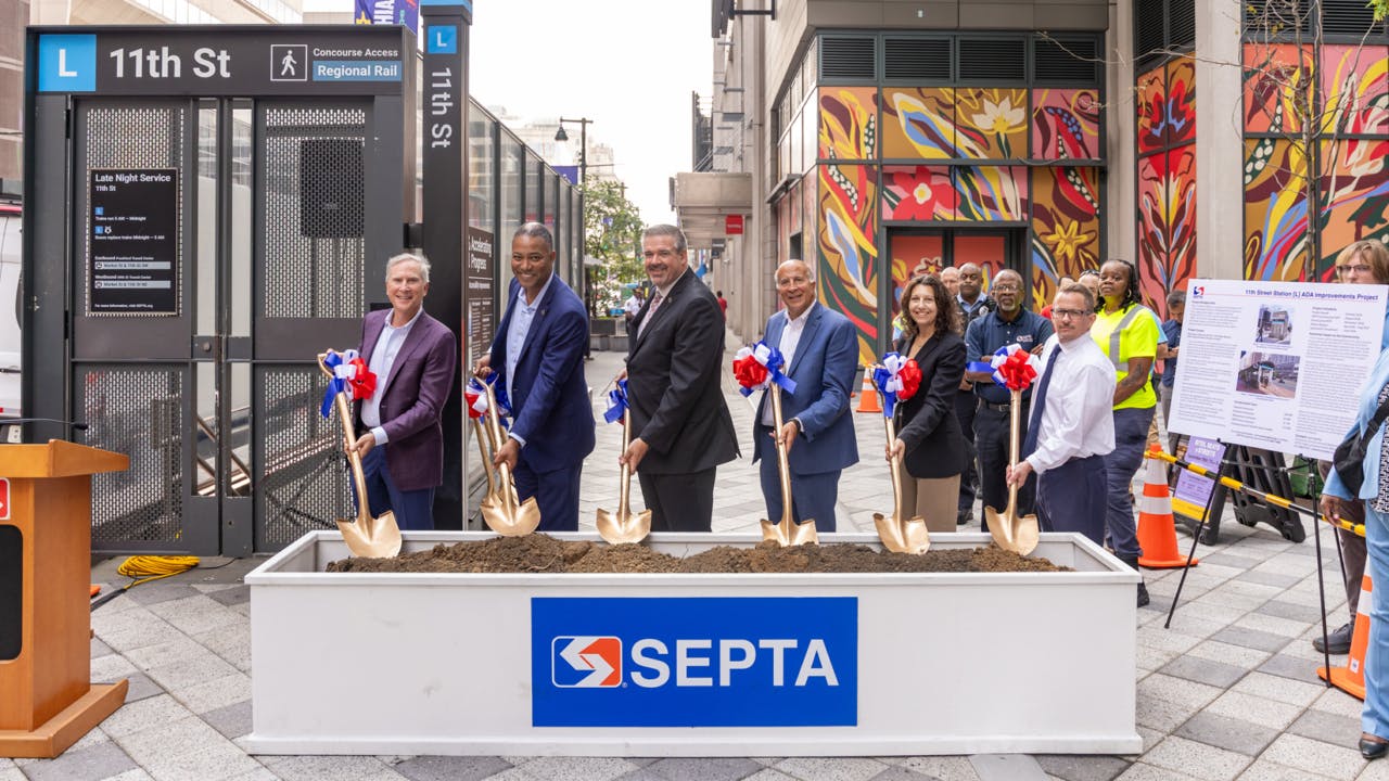 SEPTA officials pose with gold ceremonial shovels at a groundbreaking event for ADA improvements at the 11th Street 'L' station, standing behind a soil-filled planter bearing the SEPTA logo with the station entrance and a colorful mural in the background.
