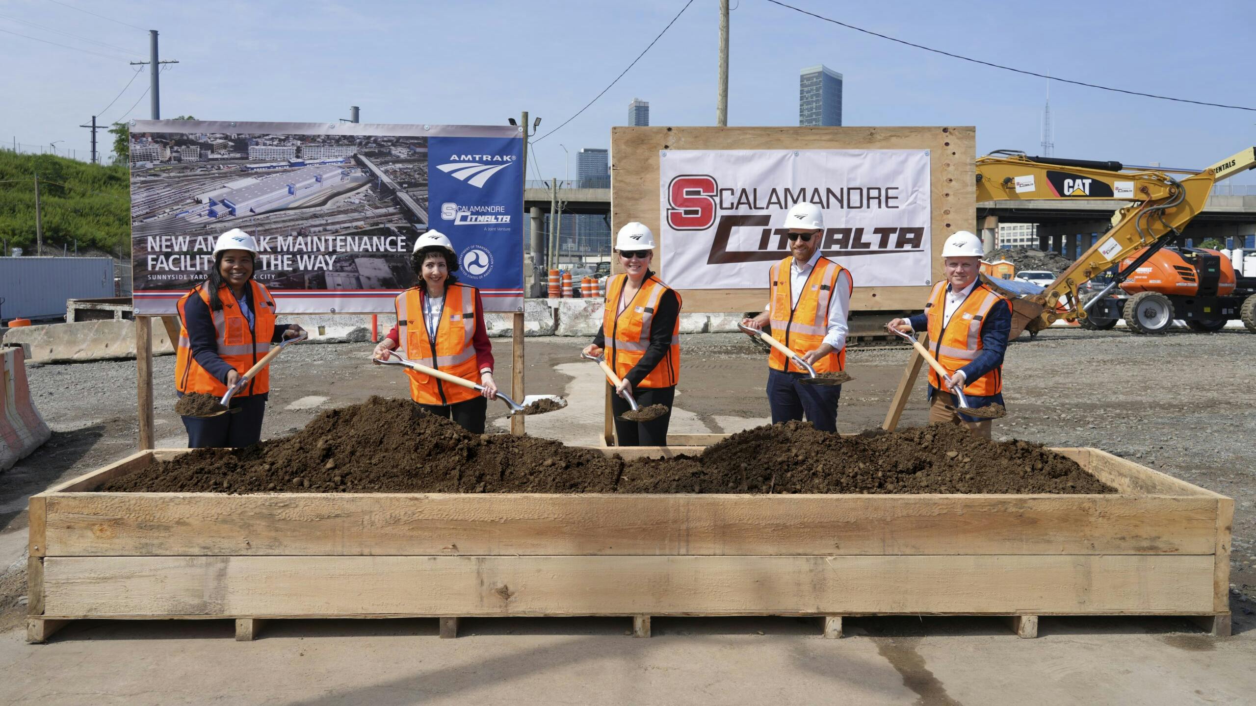 Five officials in orange safety vests and hard hats pose with shovels at a groundbreaking ceremony for Amtrak's new Sunnyside Yard maintenance facility, standing before Amtrak and Scalamandre-Citnalta joint venture signage with construction equipment and the New York City skyline in the background.