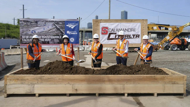 Five officials in orange safety vests and hard hats pose with shovels at a groundbreaking ceremony for Amtrak's new Sunnyside Yard maintenance facility, standing before Amtrak and Scalamandre-Citnalta joint venture signage with construction equipment and the New York City skyline in the background.