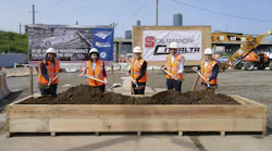 Five officials in orange safety vests and hard hats pose with shovels at a groundbreaking ceremony for Amtrak's new Sunnyside Yard maintenance facility, standing before Amtrak and Scalamandre-Citnalta joint venture signage with construction equipment and the New York City skyline in the background.