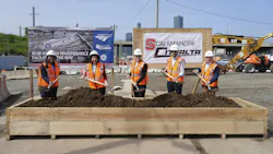 Five officials in orange safety vests and hard hats pose with shovels at a groundbreaking ceremony for Amtrak's new Sunnyside Yard maintenance facility, standing before Amtrak and Scalamandre-Citnalta joint venture signage with construction equipment and the New York City skyline in the background.