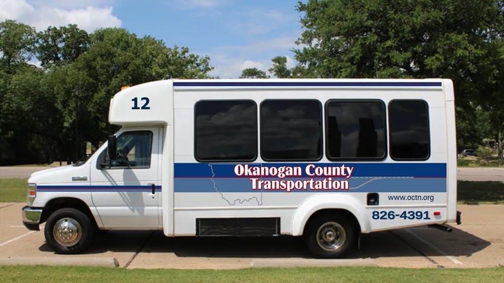 Okanogan County Transportation bus number 12, a white Ford E-Series cutaway small transit bus with a blue and red striped livery and the agency's name, website and phone number displayed on the side, parked in a lot surrounded by trees.