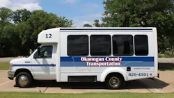 Okanogan County Transportation bus number 12, a white Ford E-Series cutaway small transit bus with a blue and red striped livery and the agency's name, website and phone number displayed on the side, parked in a lot surrounded by trees.
