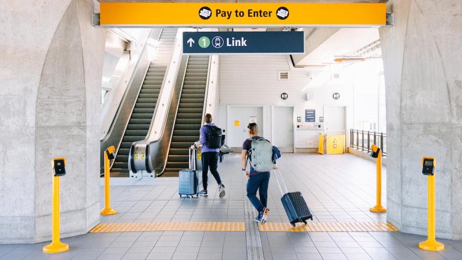 Two travelers with rolling suitcases and backpacks walk toward a Link light rail entrance at Northgate Station, passing under 'Pay to Enter' and directional signage near an escalator.