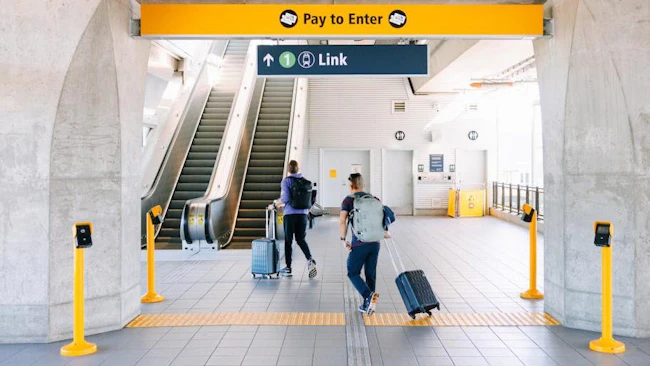 Two travelers with rolling suitcases and backpacks walk toward a Link light rail entrance at Northgate Station, passing under 'Pay to Enter' and directional signage near an escalator.