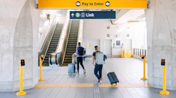 Two travelers with rolling suitcases and backpacks walk toward a Link light rail entrance at Northgate Station, passing under 'Pay to Enter' and directional signage near an escalator.