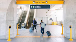Two travelers with rolling suitcases and backpacks walk toward a Link light rail entrance at Northgate Station, passing under 'Pay to Enter' and directional signage near an escalator.