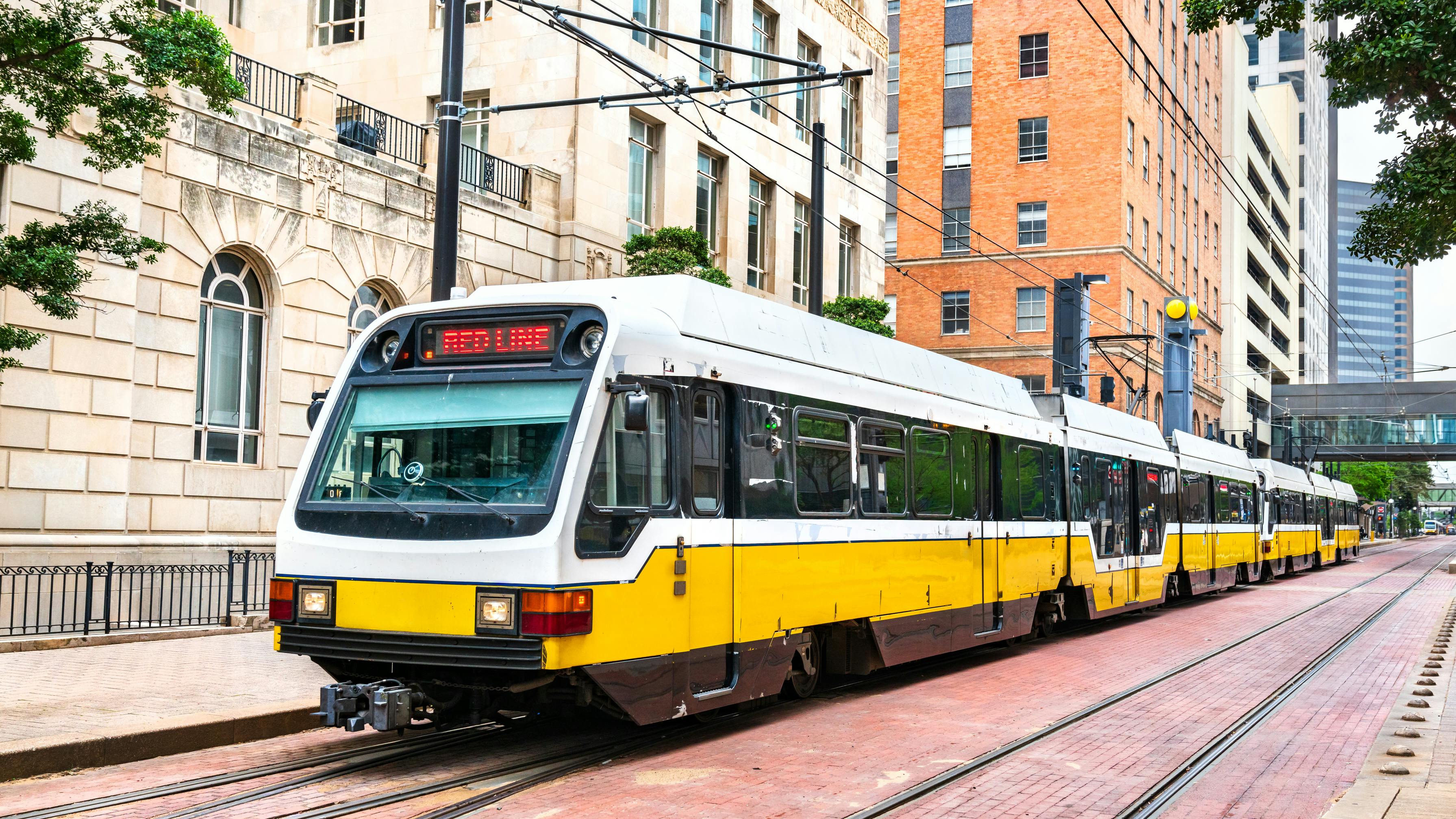 A DART Red Line light rail train, in yellow and white livery, travels on street-level tracks through downtown Dallas, framed by historic stone and brick buildings.