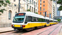 A DART Red Line light rail train, in yellow and white livery, travels on street-level tracks through downtown Dallas, framed by historic stone and brick buildings.