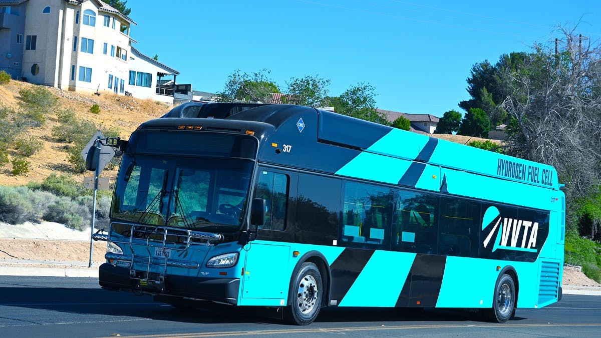 Victor Valley Transit Authority hydrogen fuel cell bus number 317 in a bold teal and black livery with 'Hydrogen Fuel Cell' lettering along the roofline navigates a desert community street in the Victor Valley area of Southern California under a clear blue sky.