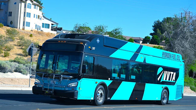 Victor Valley Transit Authority hydrogen fuel cell bus number 317 in a bold teal and black livery with 'Hydrogen Fuel Cell' lettering along the roofline navigates a desert community street in the Victor Valley area of Southern California under a clear blue sky.