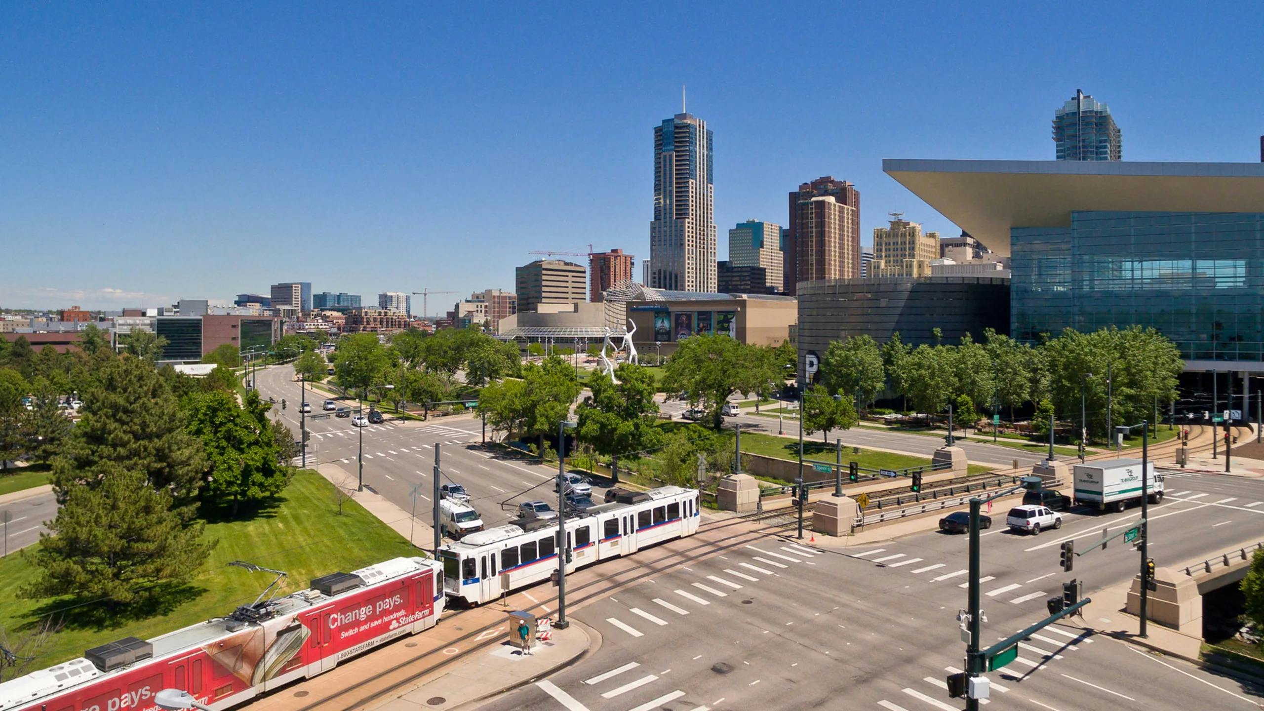 A Denver Regional Transportation District light-rail train travels along a street-level corridor near Kalamath Street in downtown Denver, Colo., with the Colorado Convention Center's distinctive blue bear sculpture and the Denver skyline visible in the background under a clear blue sky.