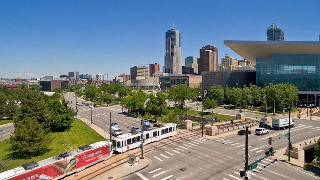 A Denver Regional Transportation District light-rail train travels along a street-level corridor near Kalamath Street in downtown Denver, Colo., with the Colorado Convention Center's distinctive blue bear sculpture and the Denver skyline visible in the background under a clear blue sky.