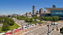 A Denver Regional Transportation District light-rail train travels along a street-level corridor near Kalamath Street in downtown Denver, Colo., with the Colorado Convention Center's distinctive blue bear sculpture and the Denver skyline visible in the background under a clear blue sky.