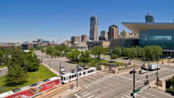 A Denver Regional Transportation District light-rail train travels along a street-level corridor near Kalamath Street in downtown Denver, Colo., with the Colorado Convention Center's distinctive blue bear sculpture and the Denver skyline visible in the background under a clear blue sky.