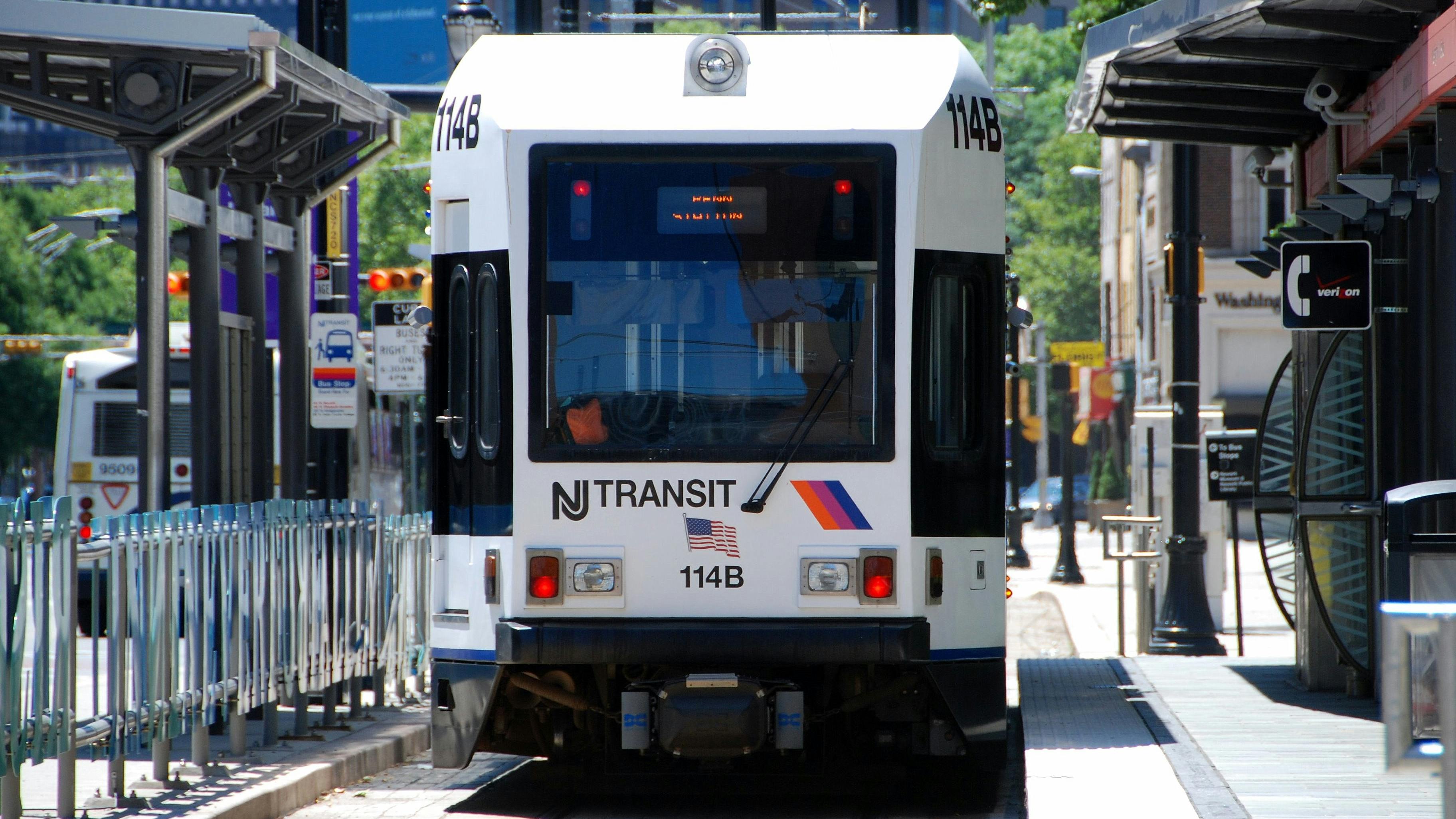 An NJ Transit light rail vehicle, numbered 114B, approaches a platform along a tree-lined city street, with a bus visible on the adjacent roadway and buildings in the background.