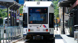 An NJ Transit light rail vehicle, numbered 114B, approaches a platform along a tree-lined city street, with a bus visible on the adjacent roadway and buildings in the background.