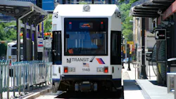 An NJ Transit light rail vehicle, numbered 114B, approaches a platform along a tree-lined city street, with a bus visible on the adjacent roadway and buildings in the background.
