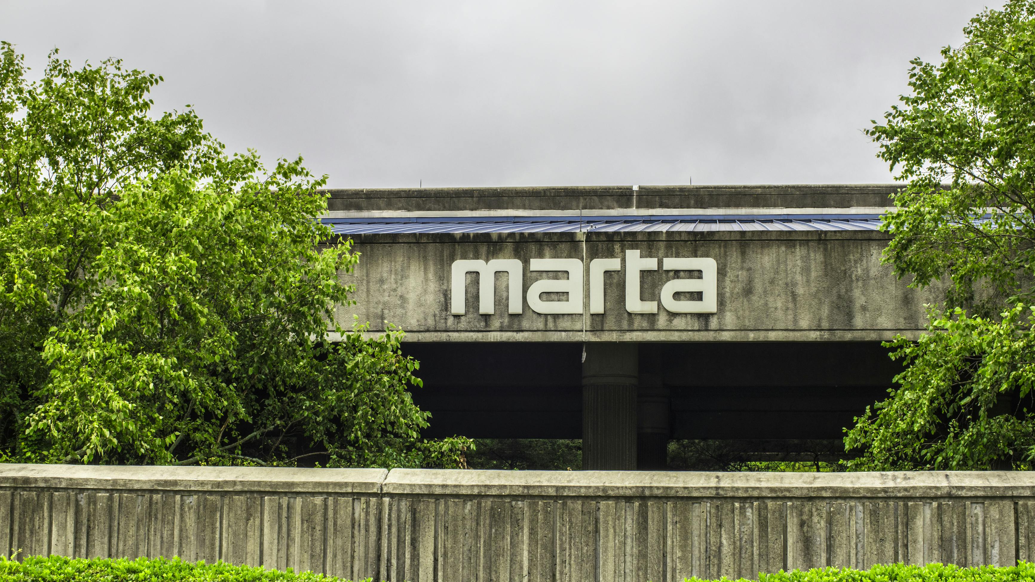 The MARTA logo mounted on the exterior of a concrete station building, framed by trees and a low retaining wall under an overcast sky.