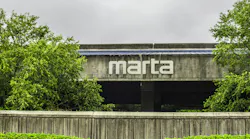 The MARTA logo mounted on the exterior of a concrete station building, framed by trees and a low retaining wall under an overcast sky.