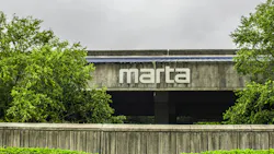 The MARTA logo mounted on the exterior of a concrete station building, framed by trees and a low retaining wall under an overcast sky.