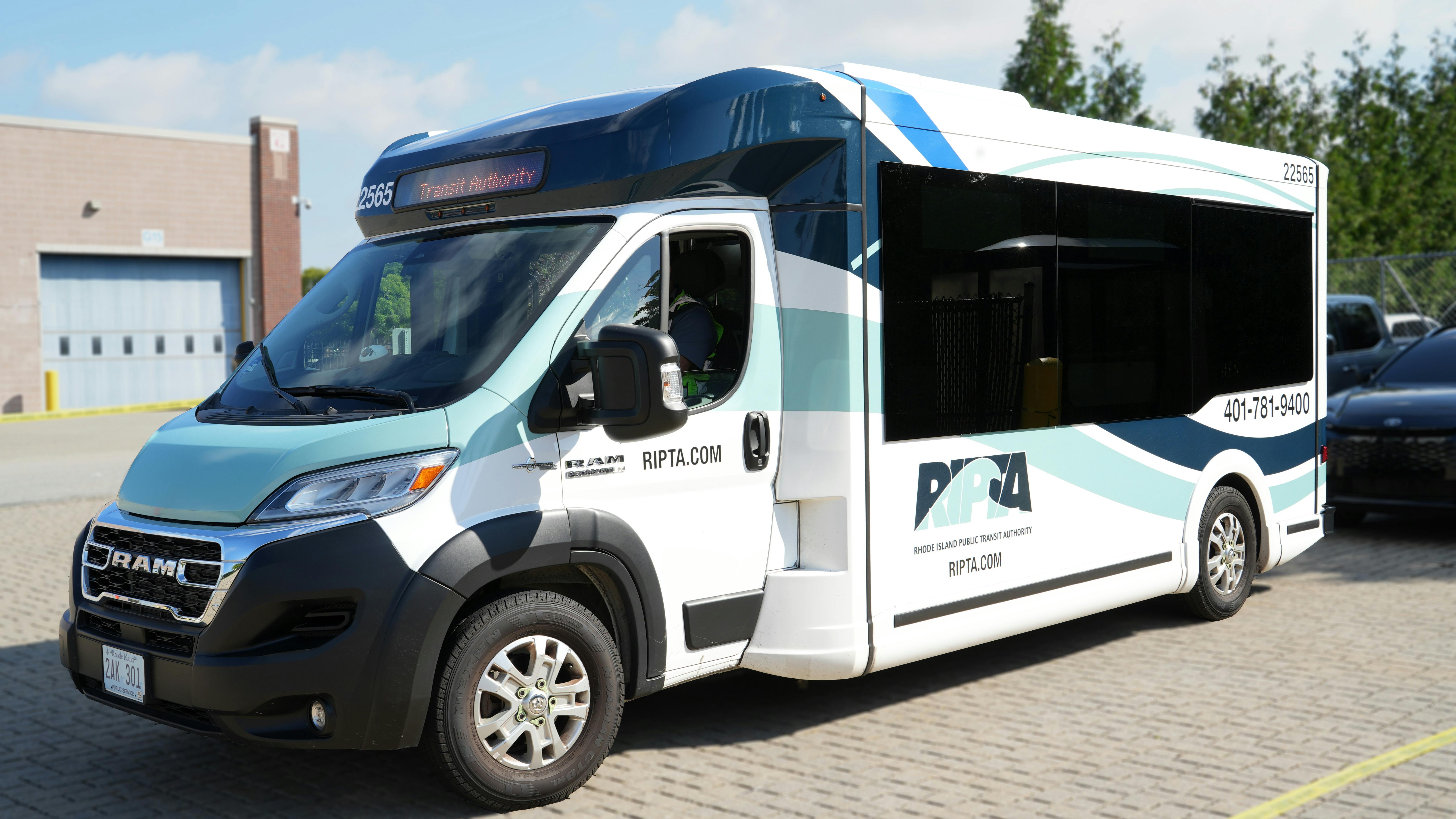 A Rhode Island Public Transit Authority (RIPTA) Model 1 Frontrunner van number 22565 in a white, navy and teal wave livery displaying 'Transit Authority' on its destination board, parked on a brick surface outside a transit facility with the RIPTA logo, RIPTA.com website and phone number visible on the side.