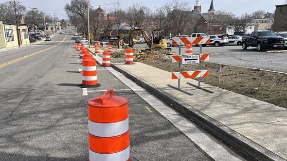 A row of orange construction barrels lines a lane of a two-lane urban street alongside an active construction zone with excavation equipment, a 'Sidewalk Closed &mdash; Cross Here' sign, and a church steeple visible in the background of a small downtown corridor.'