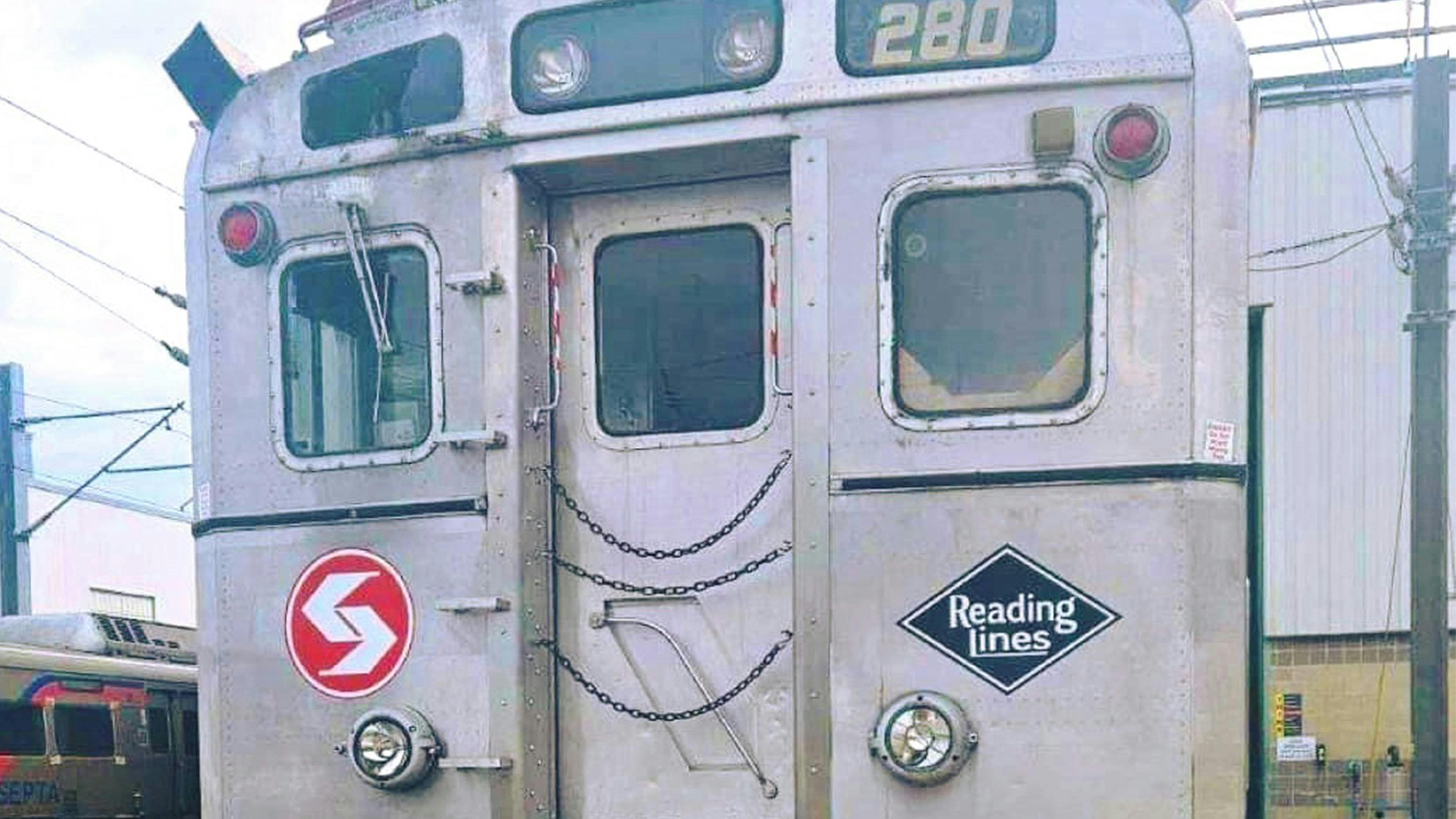 A close-up of the rear end of a retired SEPTA Silverliner commuter rail car number 280, showing the original SEPTA logo and a Reading Lines diamond emblem on its stainless steel exterior, with chain-secured doors and a newer SEPTA train visible in the background at a rail yard.