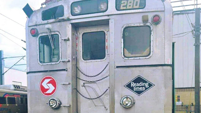 A close-up of the rear end of a retired SEPTA Silverliner commuter rail car number 280, showing the original SEPTA logo and a Reading Lines diamond emblem on its stainless steel exterior, with chain-secured doors and a newer SEPTA train visible in the background at a rail yard.