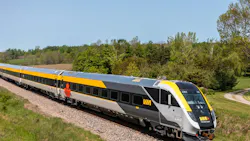 VIA Rail Canada Siemens Venture trainset number 2302 in gray and yellow livery with a Canadian maple leaf accent travels through a lush green rural landscape in Ontario on a clear spring day, with rolling farmland, trees, and a gravel country road visible in the background.