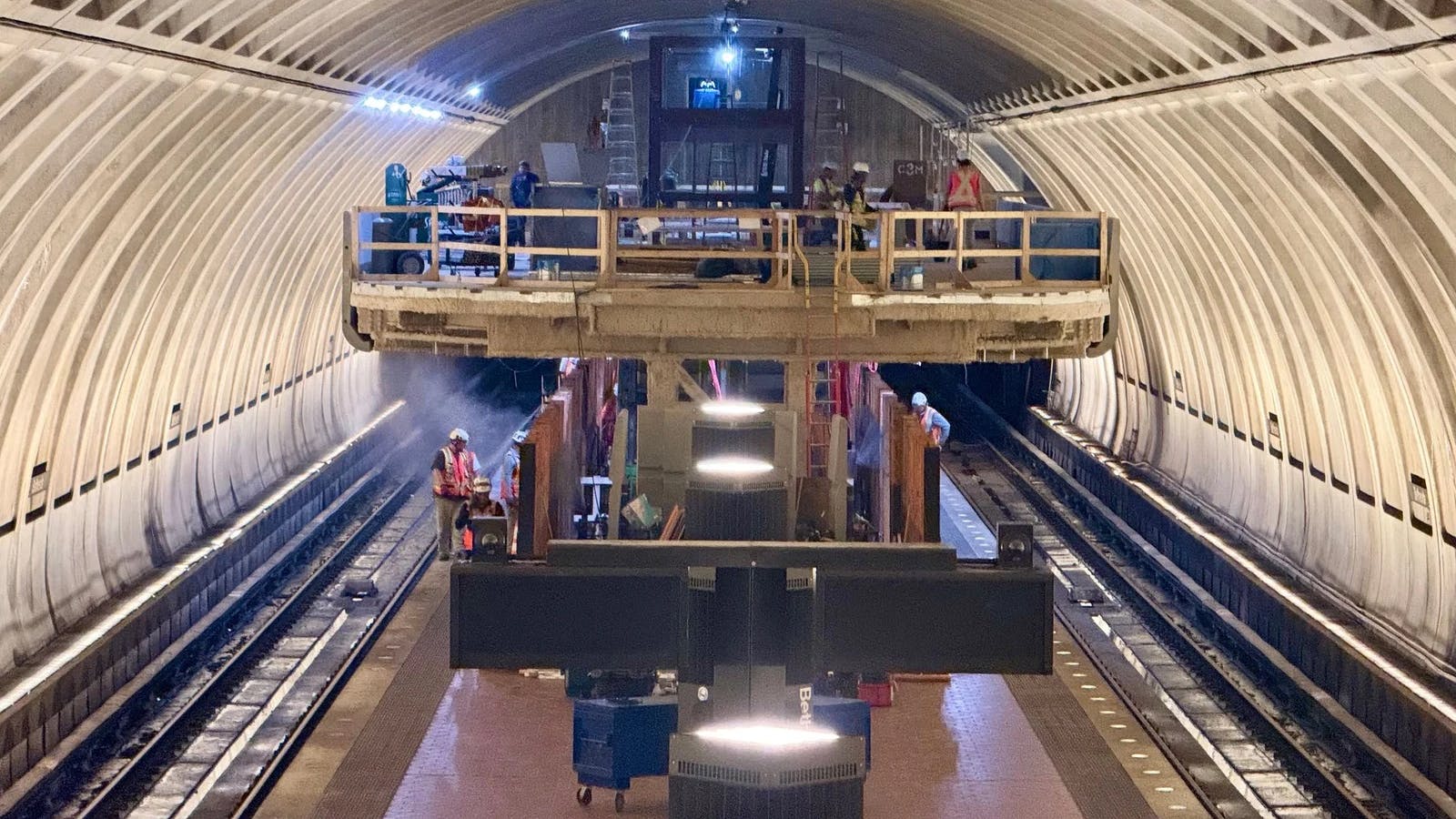 Construction workers install equipment inside an arched subway tunnel under construction, standing on a raised platform and working track-level amid staging materials and lighting rigs.