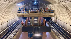 Construction workers install equipment inside an arched subway tunnel under construction, standing on a raised platform and working track-level amid staging materials and lighting rigs.
