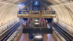 Construction workers install equipment inside an arched subway tunnel under construction, standing on a raised platform and working track-level amid staging materials and lighting rigs.