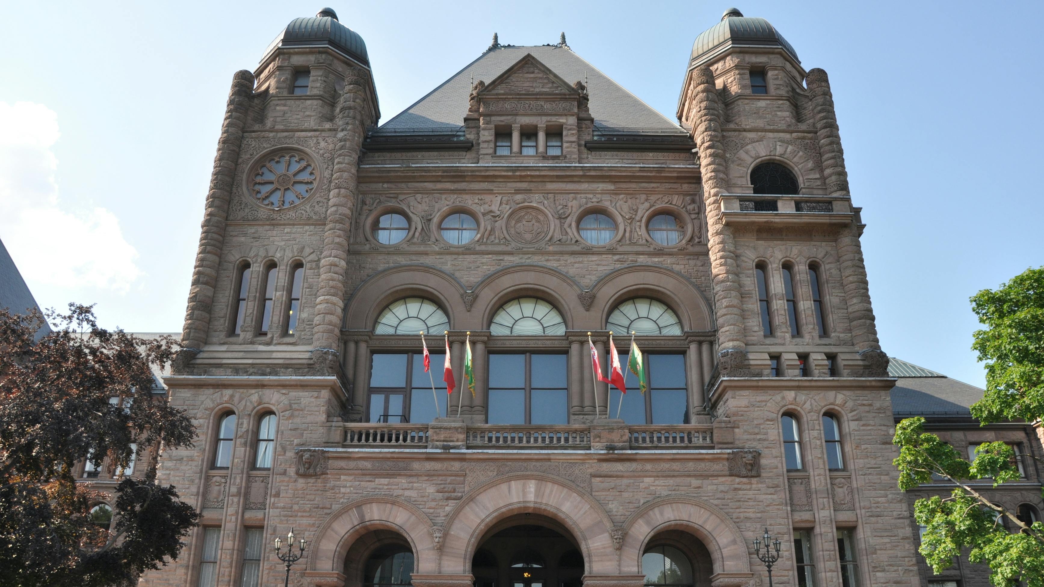 The Ontario Legislative Building at Queen's Park in Toronto, showing its ornate sandstone facade with twin towers, arched entrances, and a row of Canadian and provincial flags, framed by trees on either side.