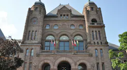 The Ontario Legislative Building at Queen's Park in Toronto, showing its ornate sandstone facade with twin towers, arched entrances, and a row of Canadian and provincial flags, framed by trees on either side.