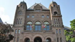 The Ontario Legislative Building at Queen's Park in Toronto, showing its ornate sandstone facade with twin towers, arched entrances, and a row of Canadian and provincial flags, framed by trees on either side.