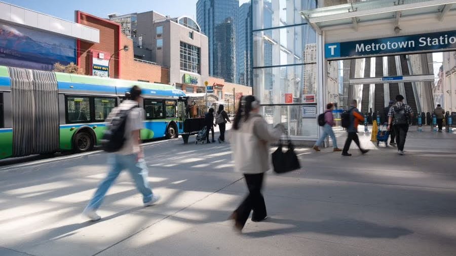 Pedestrians cross a plaza outside Metrotown Station in Burnaby, with a TransLink articulated bus passing behind them and the SkyTrain platform's escalators visible under a glass canopy, surrounded by high-rise buildings.